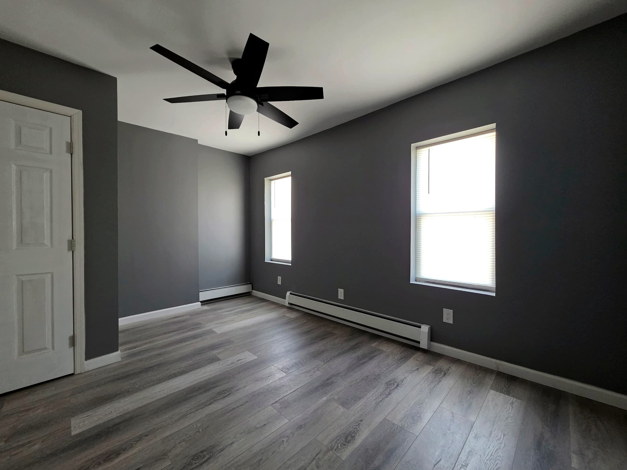 Empty living room from the corner, with two windows and a white door.