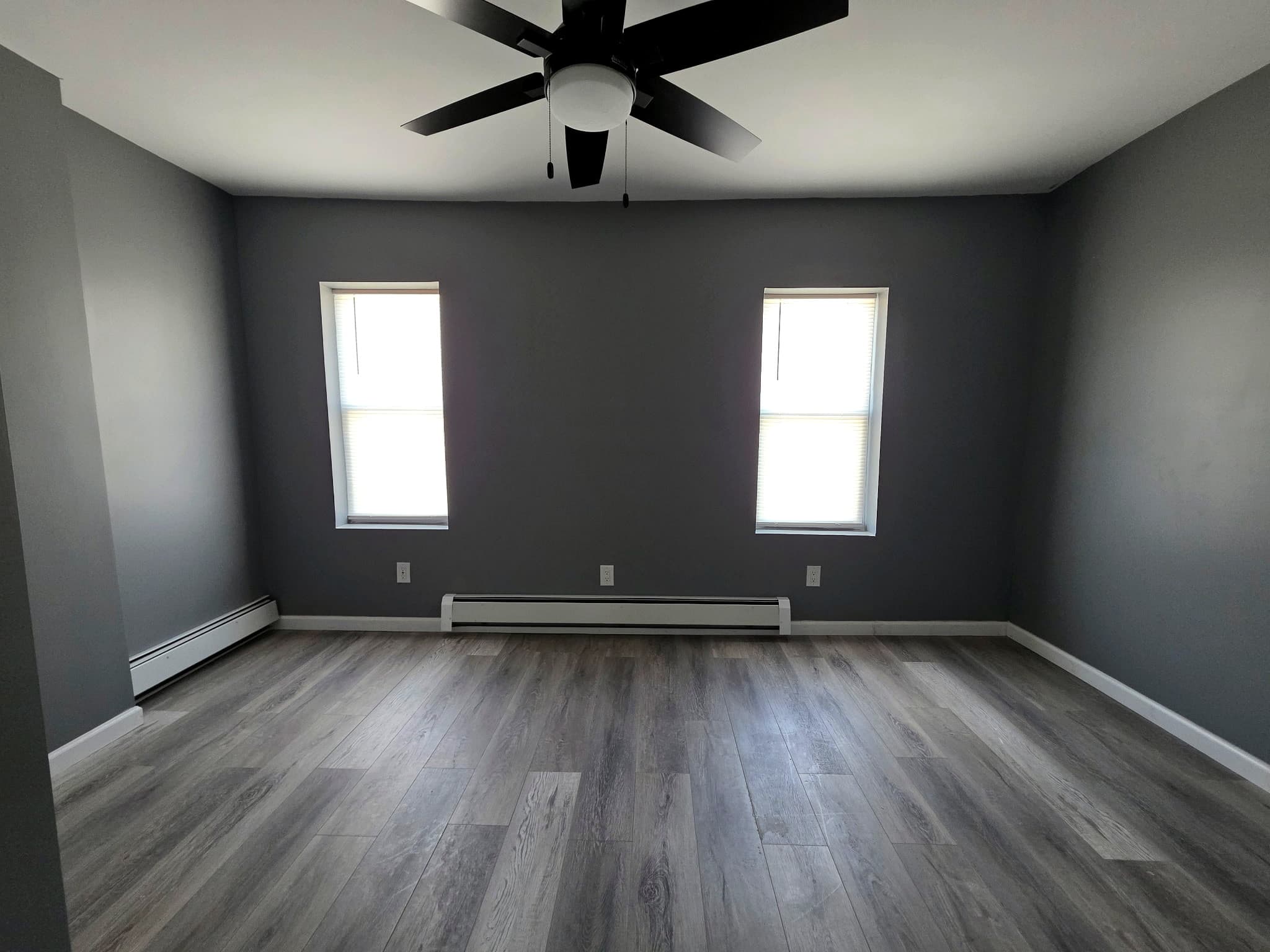 Empty living room facing two windows, with a ceiling fan and baseboard heat.